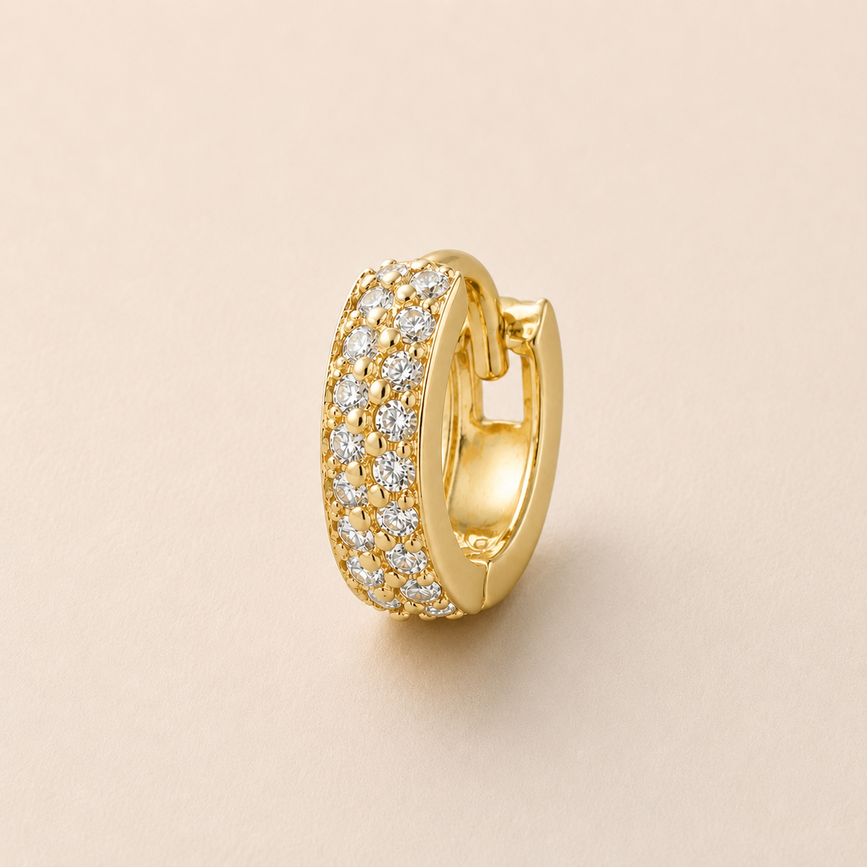 A gold huggie hoop earring paved with small clear stones, shown on a plain beige background.