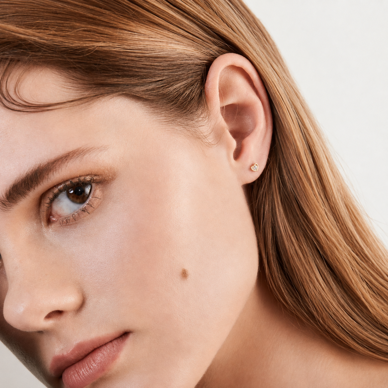Close-up of a woman's ear wearing a small, delicate gold stud earring against a clean, light background.