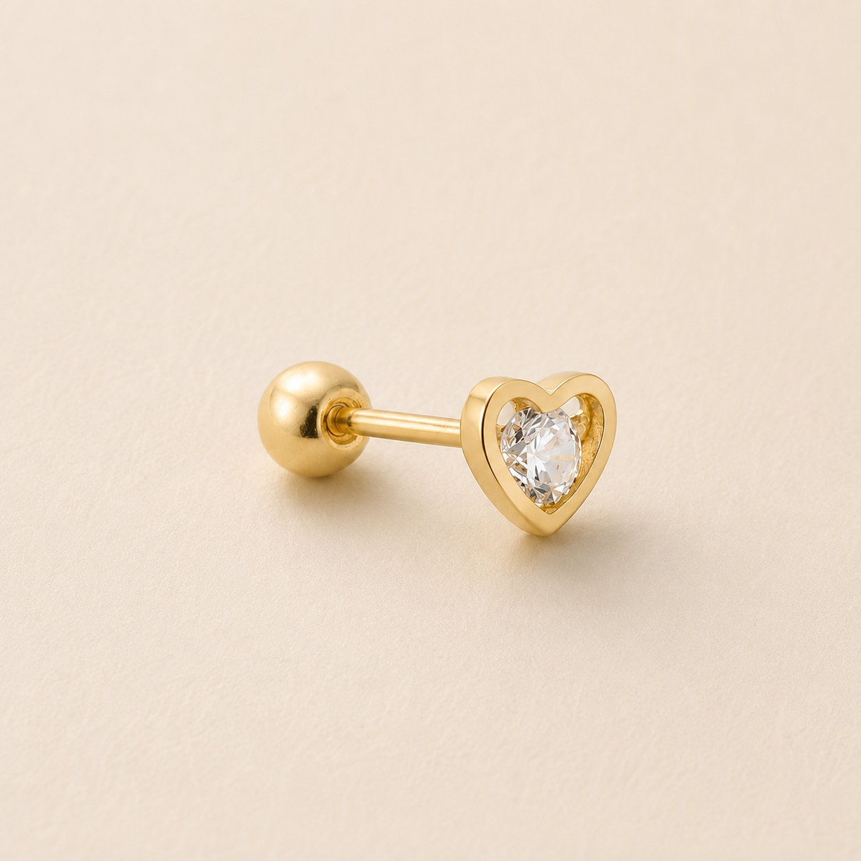 A gold-plated labret stud earring featuring a heart-shaped bezel setting with a clear heart-shaped gemstone, shown on a warm beige background.