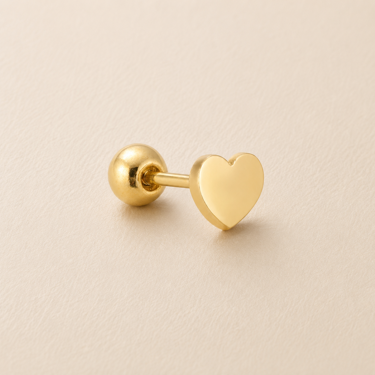 A single gold-colored stud earring featuring a flat, polished heart-shaped front and a spherical ball back, displayed on a textured beige surface.