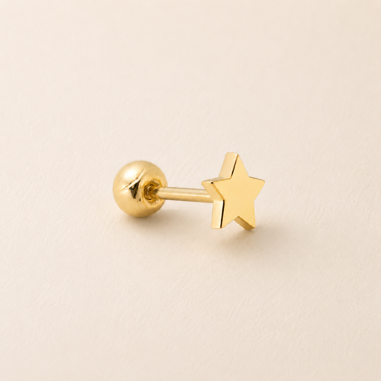 A single gold-colored labret stud earring featuring a polished star-shaped front and a round ball back, displayed on a clean, light beige textured surface.
