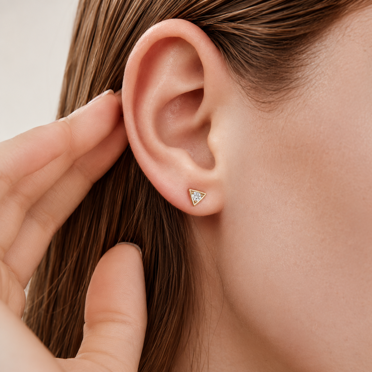 A close-up of a model's ear wearing a small triangular stud earring with a gold-toned metal setting and clear stones, with her fingers touching her hair nearby.