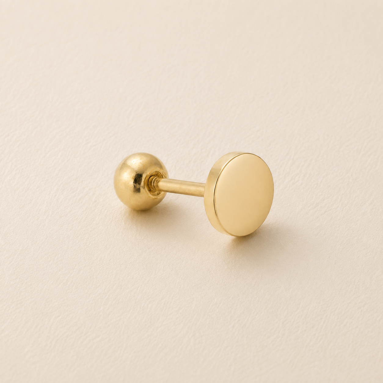 A single gold-colored stud earring with a flat circular disc front and a spherical ball back, resting on a textured light beige surface.