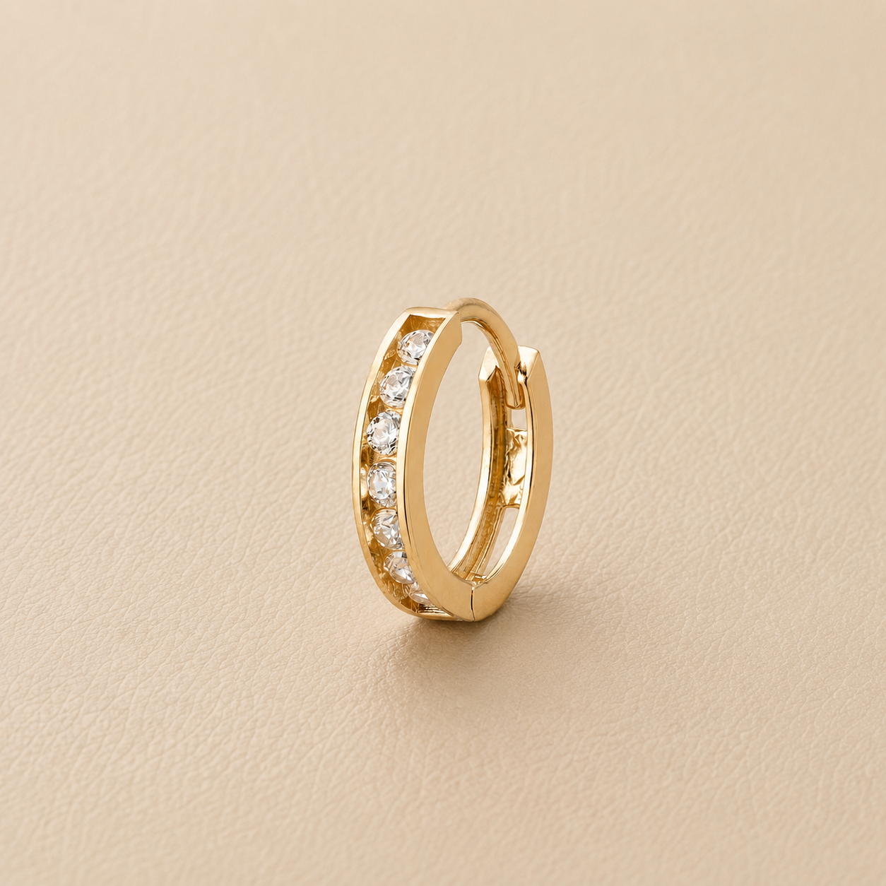 A gold huggie hoop earring with a row of clear pavé-set stones, photographed on a textured beige background.