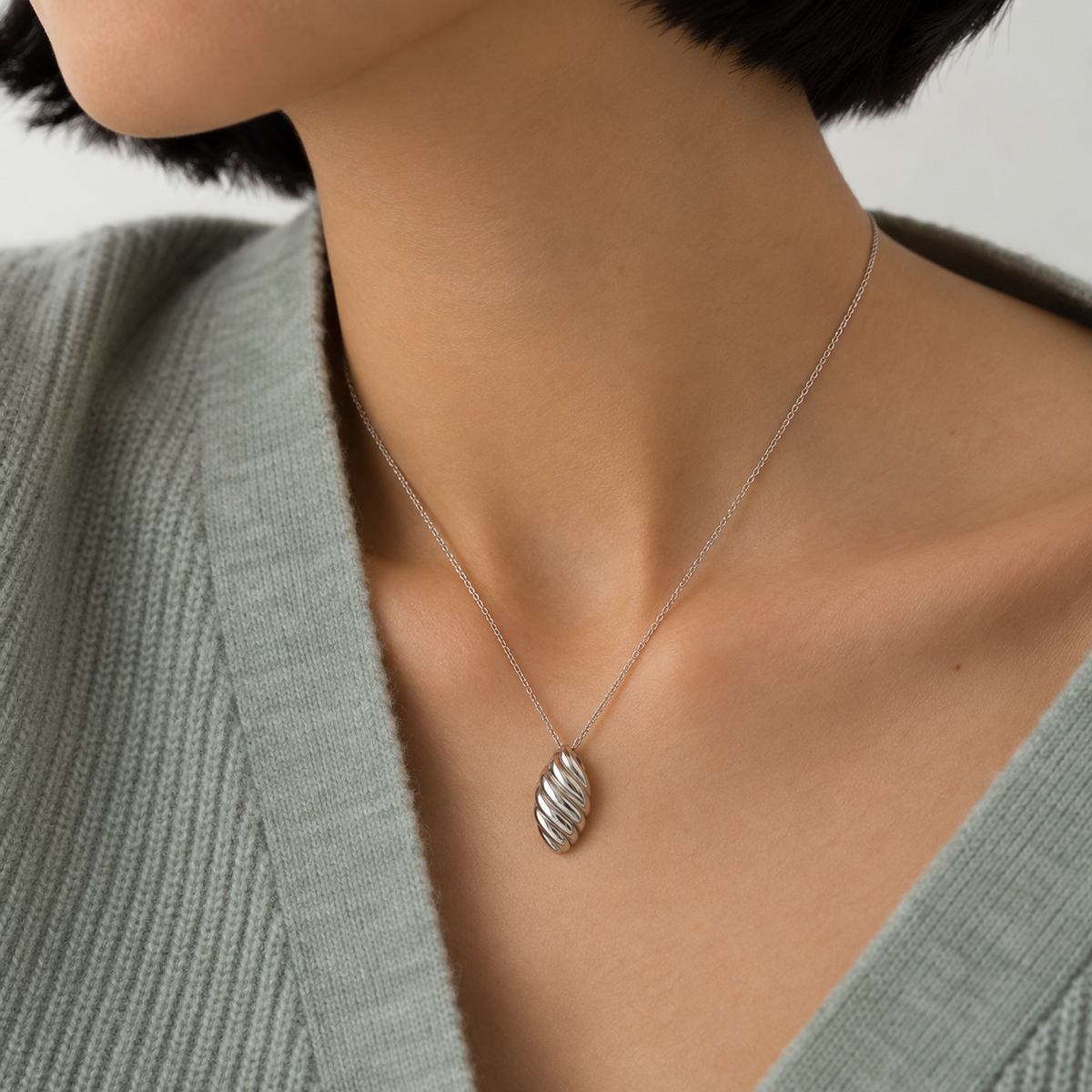 A close-up of a person wearing a delicate silver chain necklace with a textured, oval-shaped silver pendant, paired with a sage green knit top.