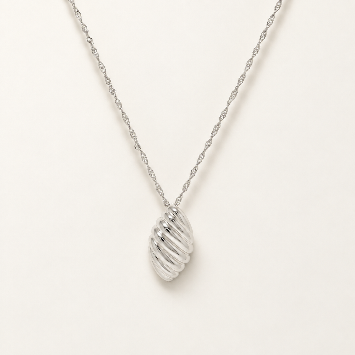 A silver pendant necklace featuring a textured, ribbed oval drop on a delicate twisted chain, photographed against a clean off-white background.