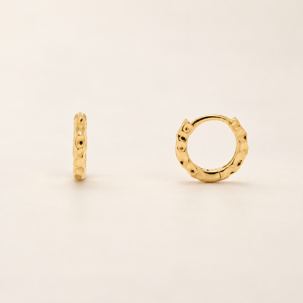 A pair of small gold huggie hoop earrings with a textured, dimpled surface, shown on a clean off-white background.