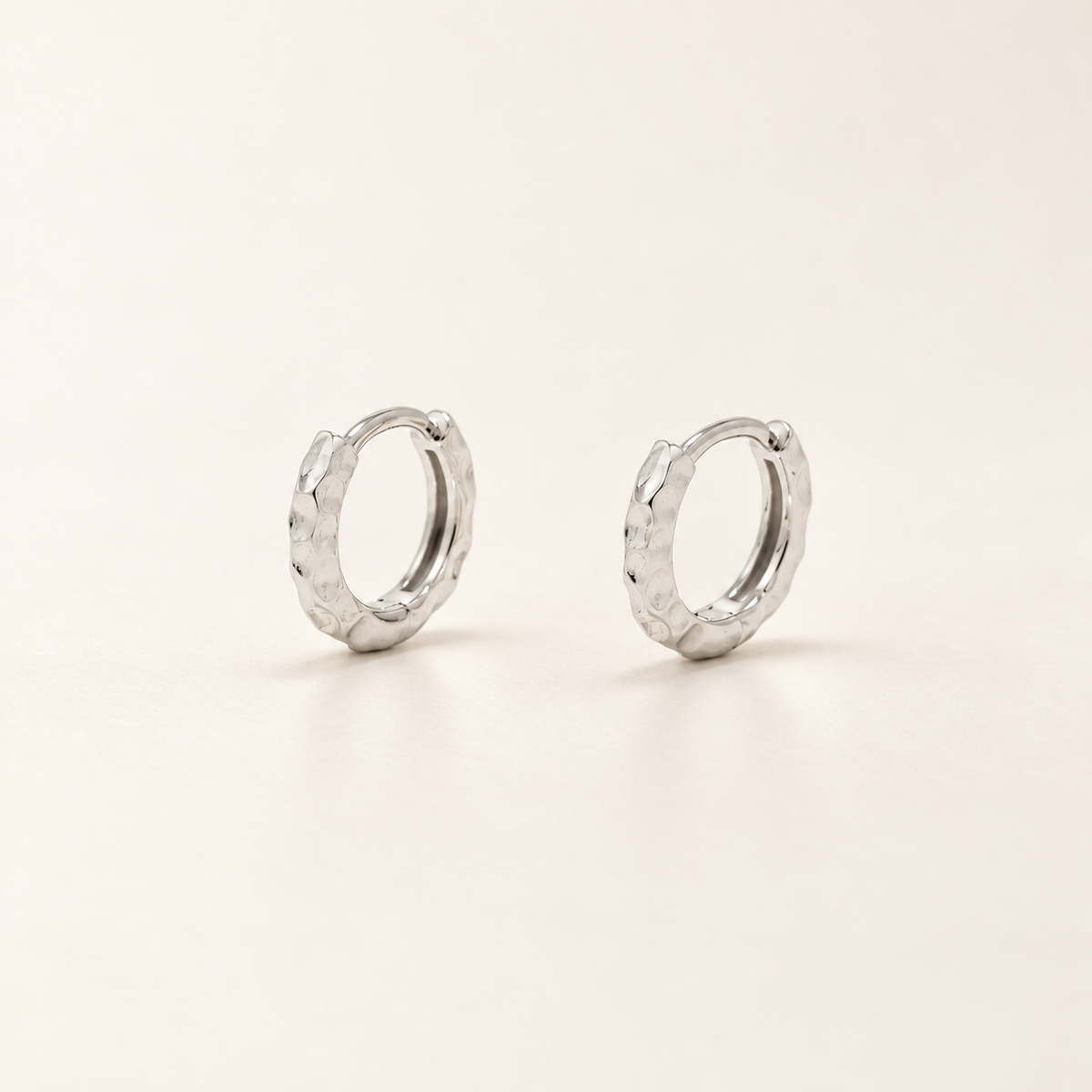 A pair of small silver huggie hoop earrings with a textured, hammered finish, displayed on a clean off-white background.
