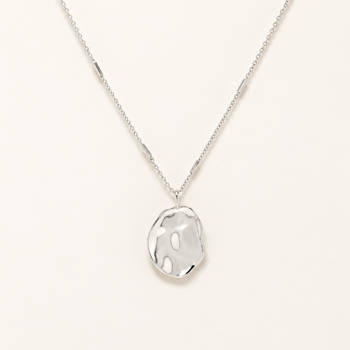 A silver necklace with a polished, organic-shaped pendant on a delicate chain, presented against a clean, off-white background.