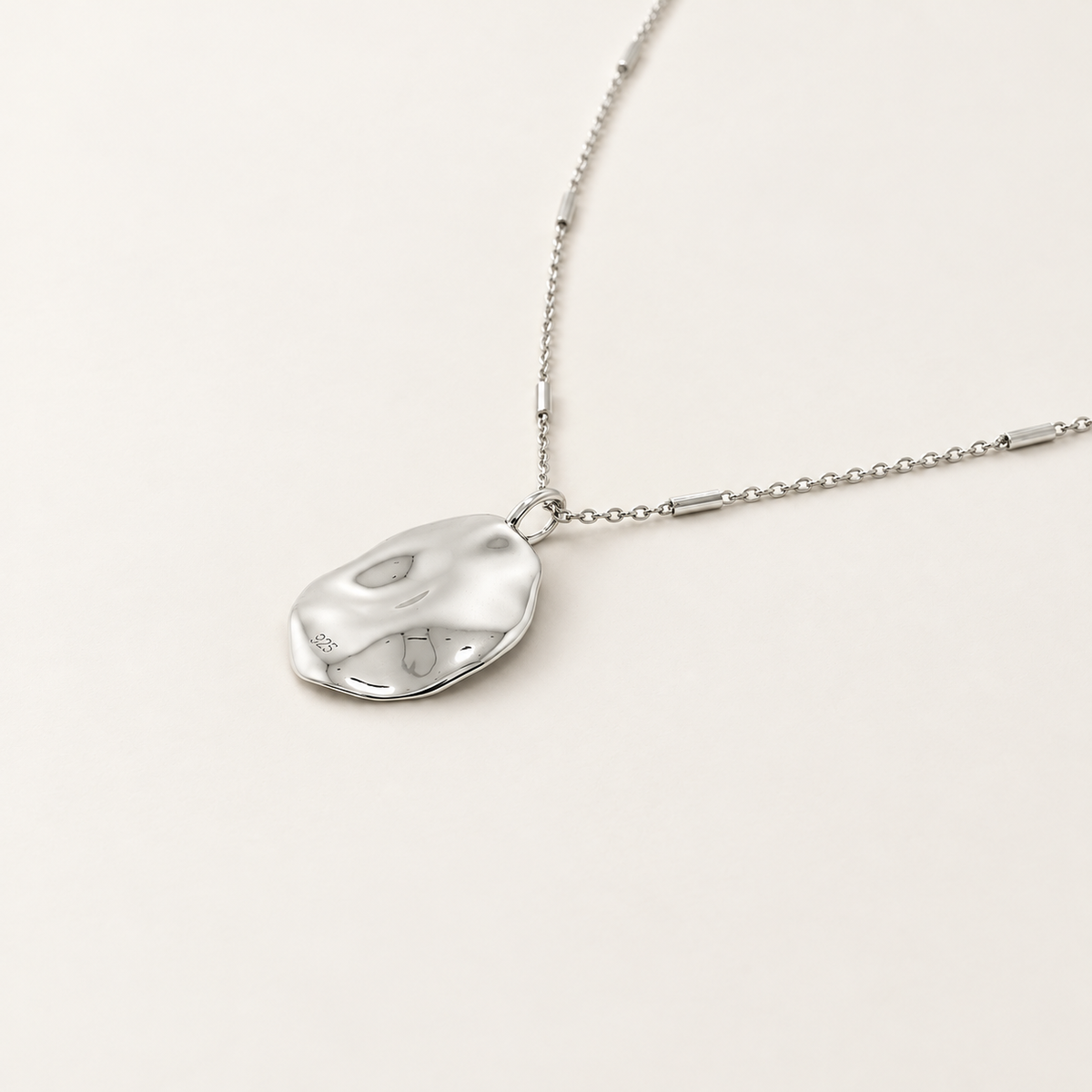 A polished silver pendant necklace with an organic, irregularly shaped disc pendant on a delicate chain, displayed on a clean, light beige background.