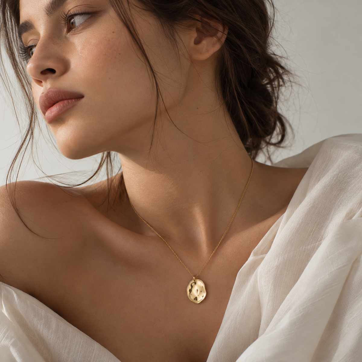 A close-up of a woman wearing a delicate gold necklace with a small, textured gold pendant, styled with a white draped top against a soft neutral background.