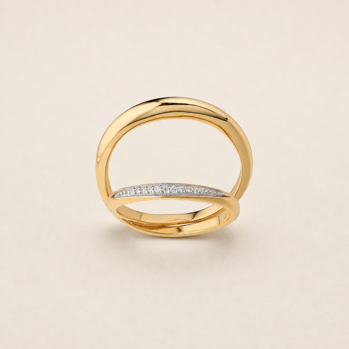A gold ring with a polished, open double-band design and a curved row of small clear stones across the front, set against a light beige background.