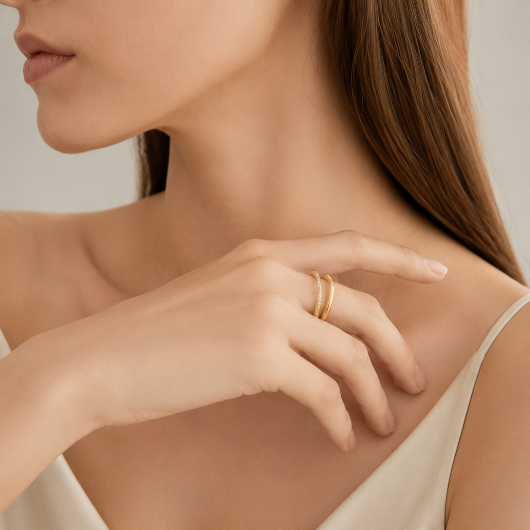 A close-up of a woman wearing delicate gold rings on her finger, wearing a cream-colored satin top against a soft neutral background.