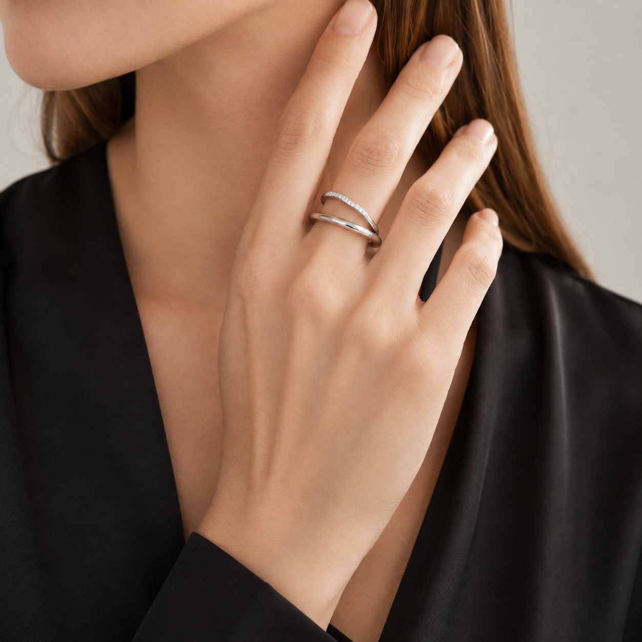 A close-up of a woman's hand wearing two slim silver rings, with her hand resting near her neck and her black satin blouse visible against a soft light gray background.