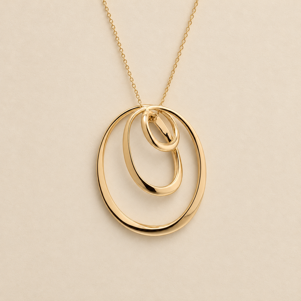 A gold pendant necklace featuring three interlocking, overlapping oval rings on a delicate chain, set against a solid light beige background.