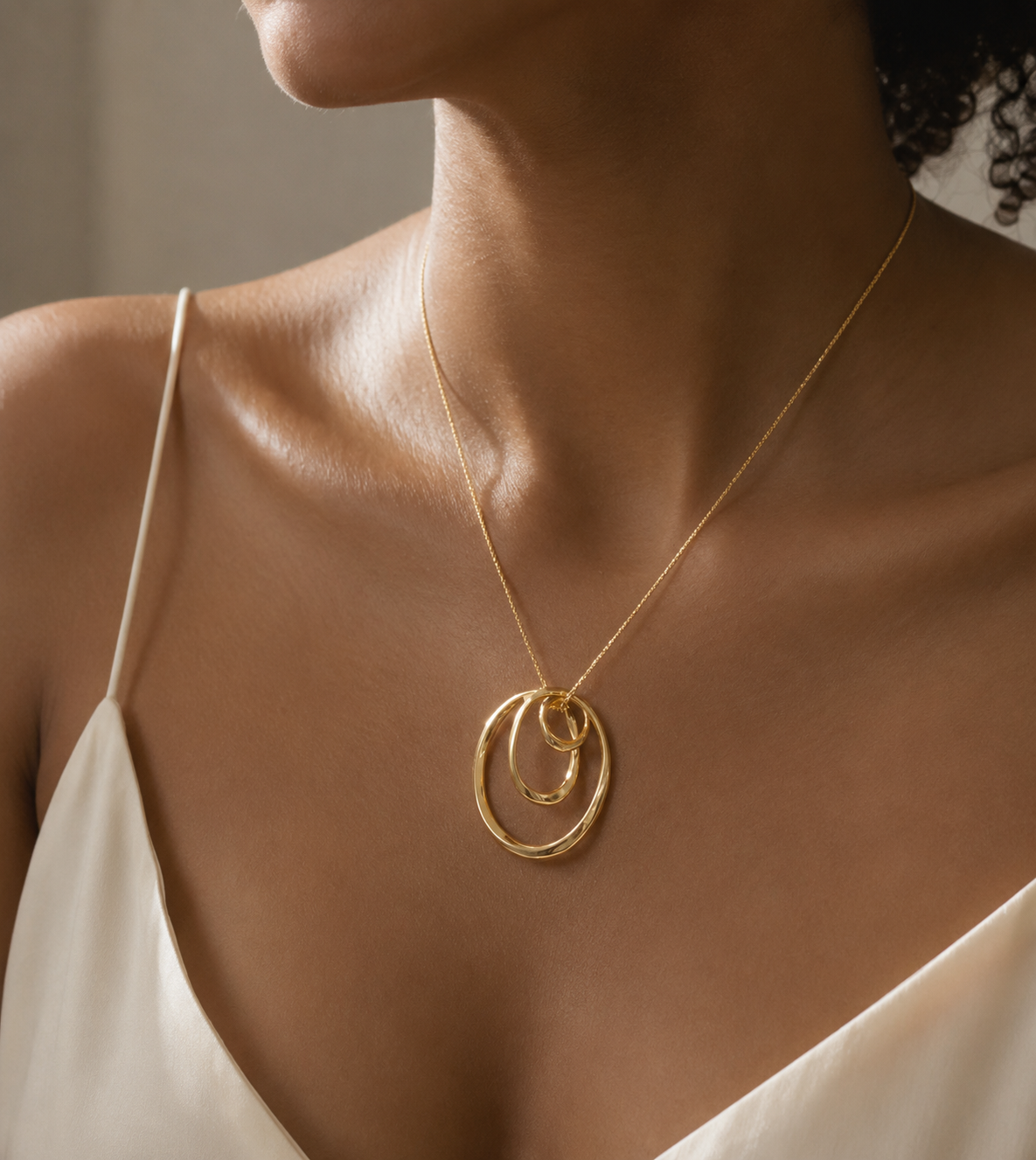 A close-up of a model wearing a delicate gold chain necklace with a double interlocking ring pendant, styled with a white camisole.