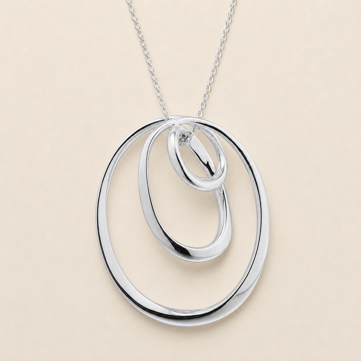 A silver pendant necklace featuring three interlocking, nested oval rings on a delicate silver chain, set against a solid light beige background.