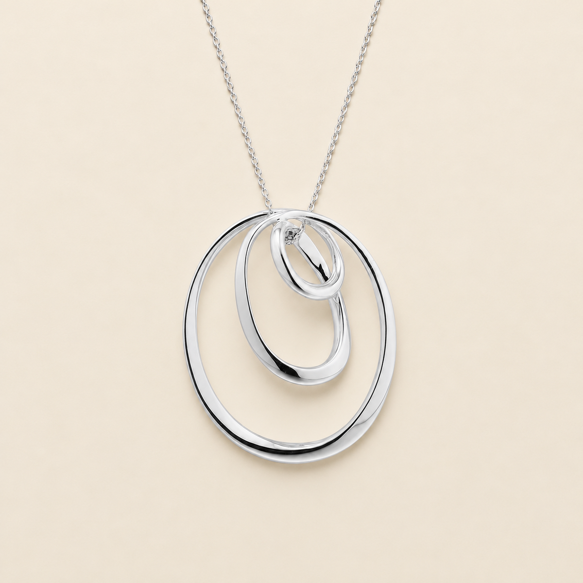 A silver pendant necklace featuring three interlocking, nested oval rings on a delicate silver chain, set against a solid light beige background.