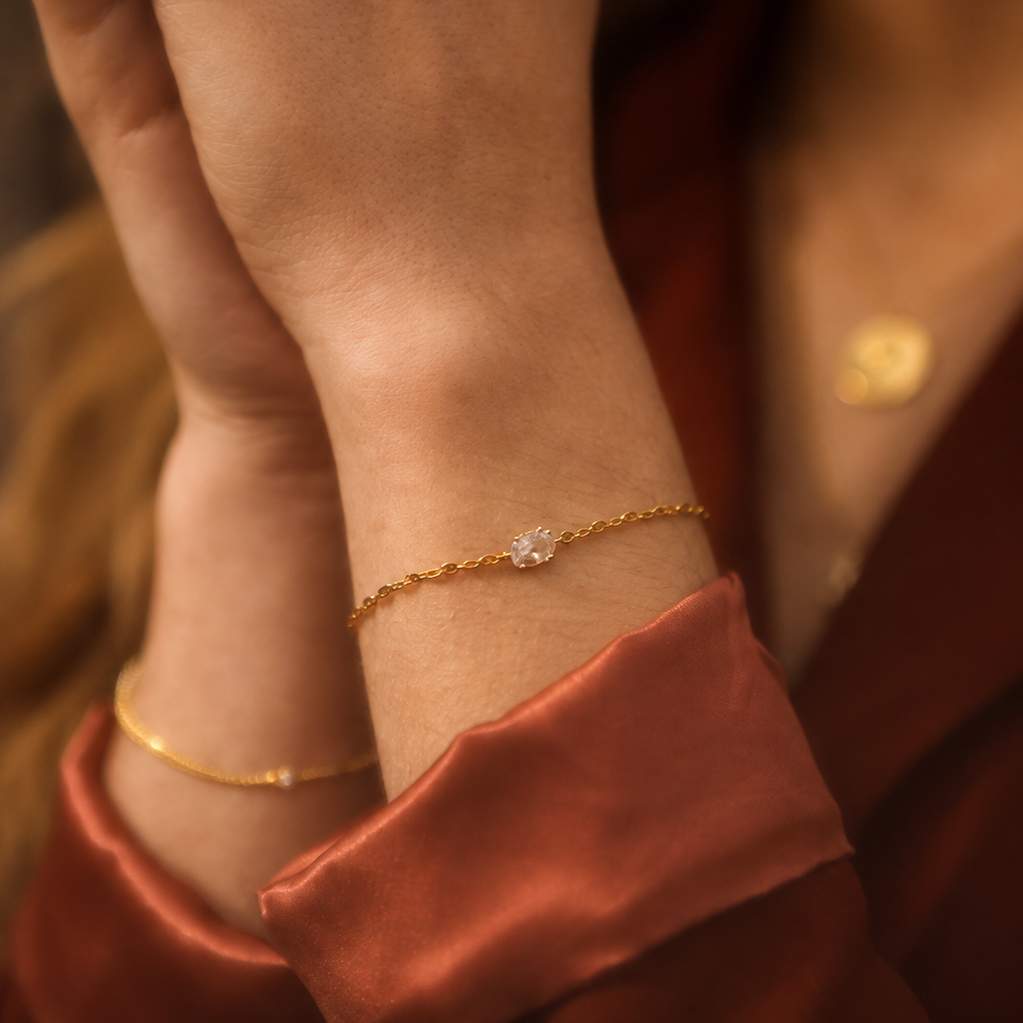 A close-up lifestyle shot of a delicate gold chain bracelet with a small clear crystal charm worn on a woman's wrist, paired with a matching gold bangle on her other wrist. She is wearing a rust-colored satin top and gold rings on her fingers.