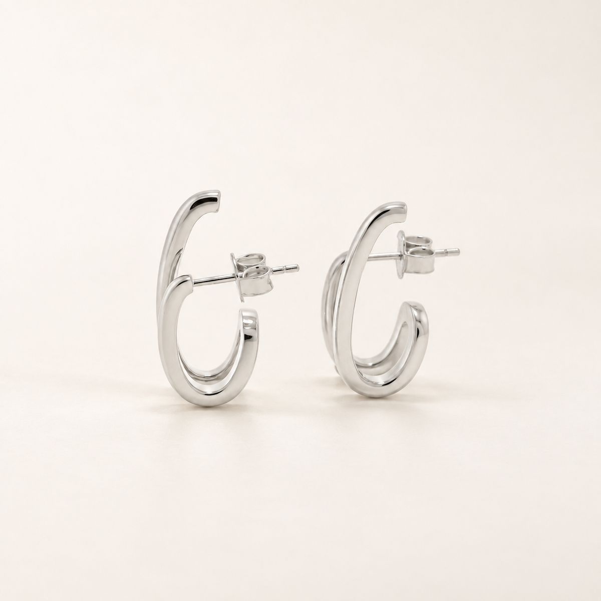 A pair of polished silver hoop earrings with a thick, curved double-band design, shown from a side profile on a clean, light beige background.