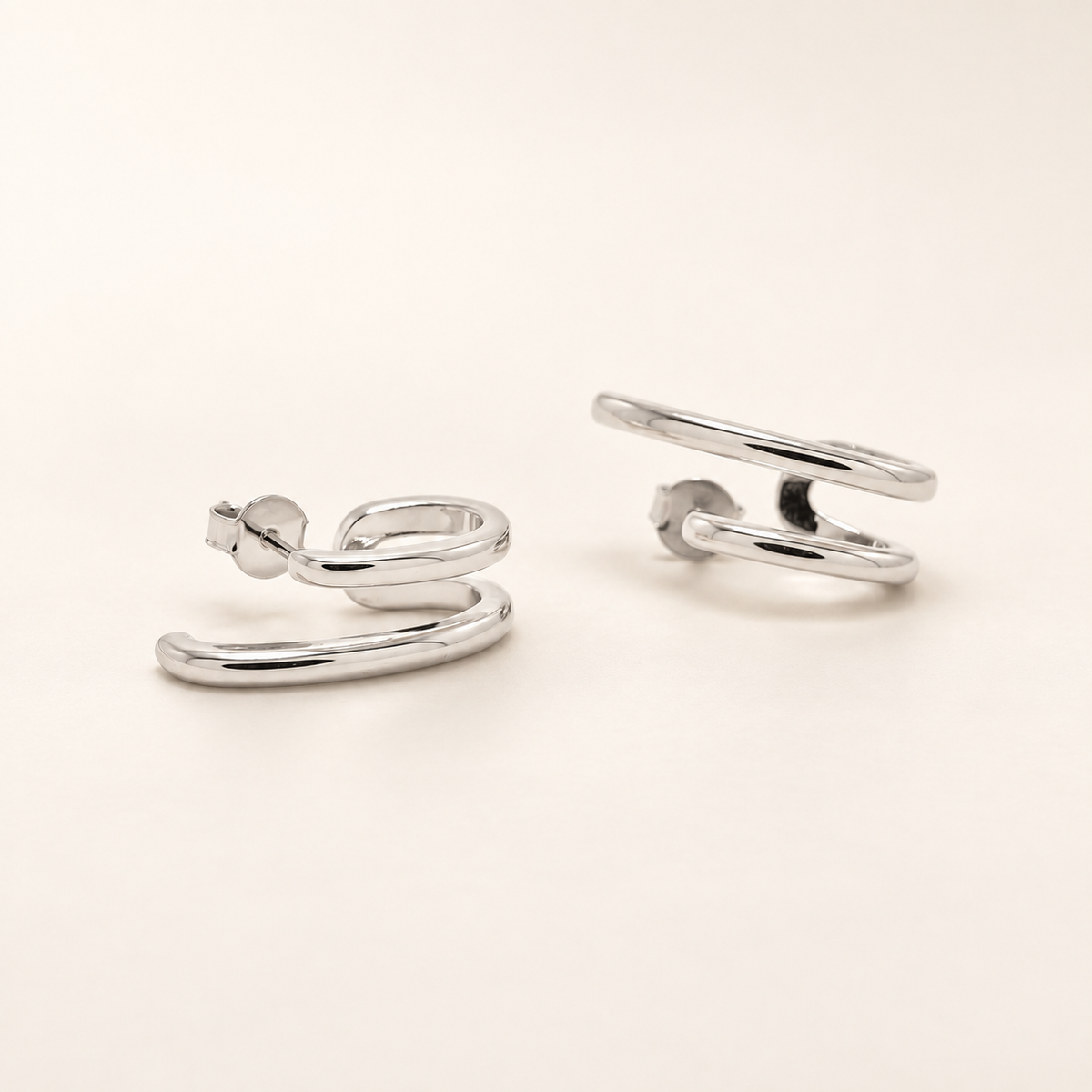 A pair of polished silver hoop earrings with a unique open spiral design, displayed on a clean, solid off-white background.