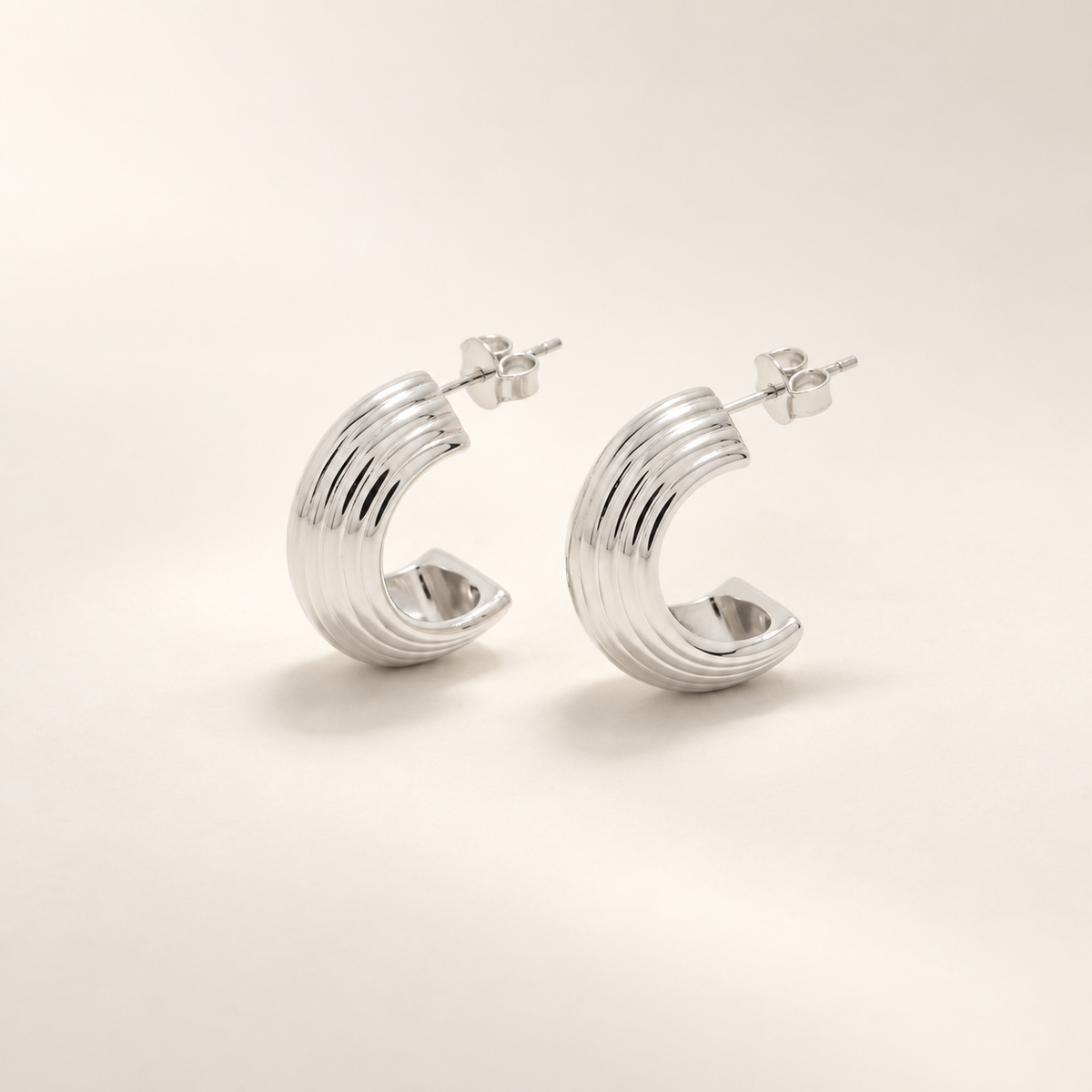 A pair of polished silver crescent-shaped hoop earrings with a ribbed, textured exterior and flat post-back closures, displayed on a clean, light beige background.