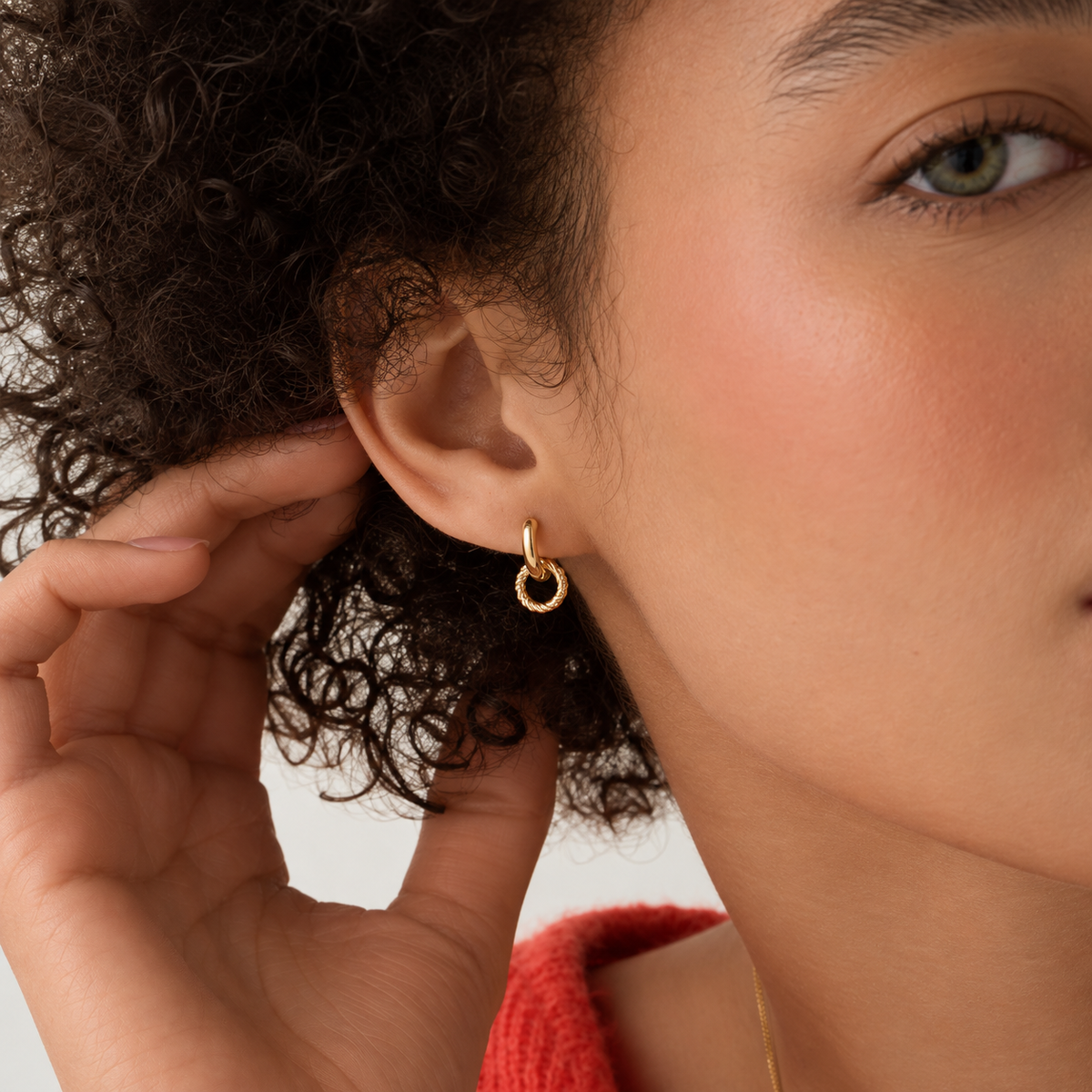 A close-up of a model wearing a small gold hoop earring with a textured, beaded inner ring. The shot focuses on the ear and earring, with the model's curly dark hair, hand, and skin visible against a clean, light background.