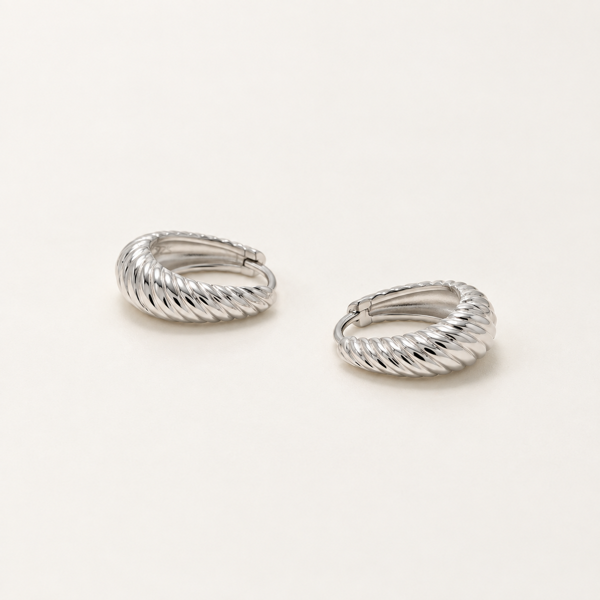 A pair of silver huggie hoop earrings with a textured, twisted rope-like band, displayed on a clean off-white background.