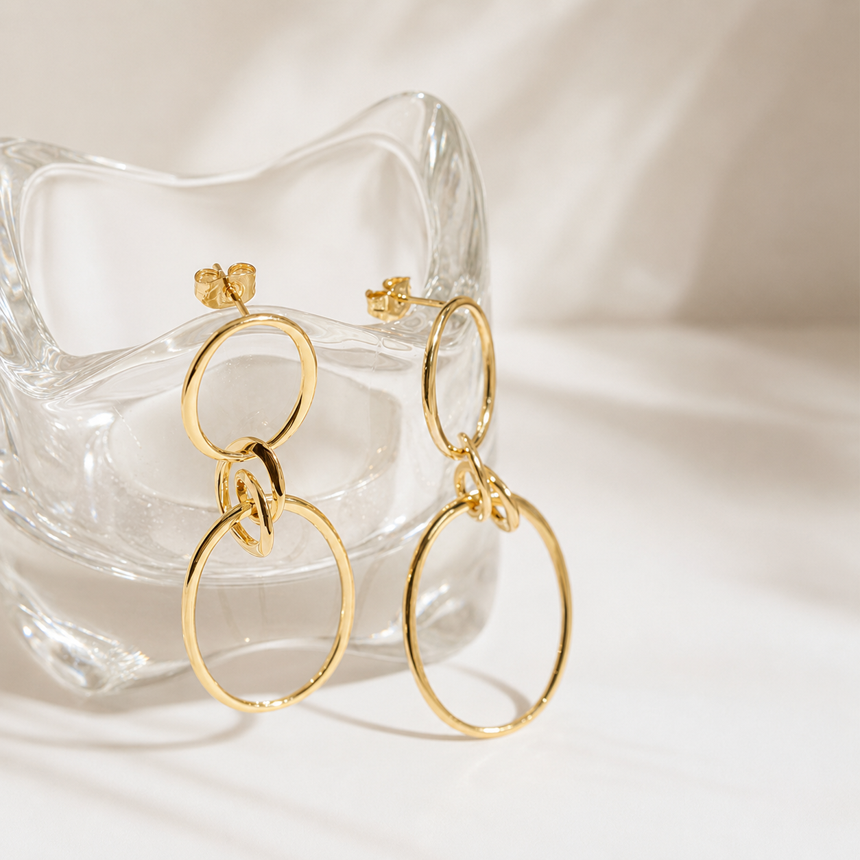 Gold hoop drop earrings with a textured knot detail, displayed on a clear glass dish against a soft white background.