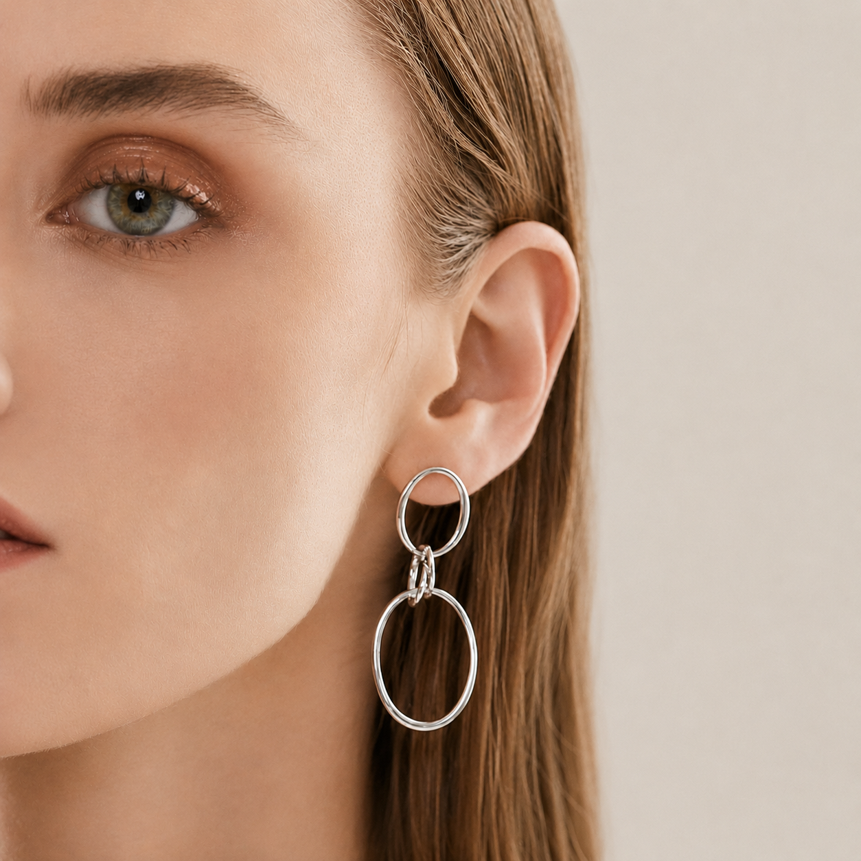 A close-up of a woman's ear wearing a silver drop earring with two interlocking oval rings, against a soft beige background.