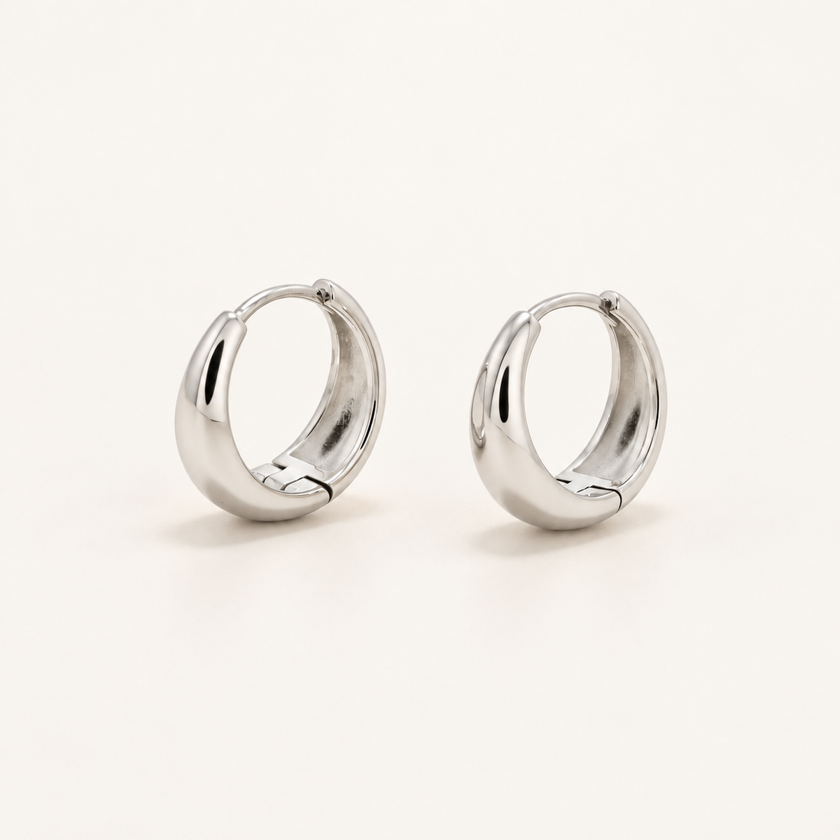 A pair of polished silver huggie hoop earrings on a clean, light off-white background.