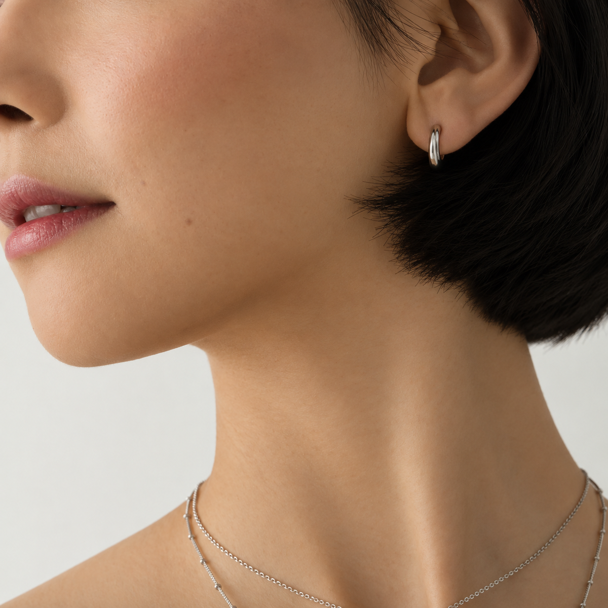 Close-up of a model's ear wearing a small silver crescent-shaped hoop earring, with part of the face, neck, and short dark hair visible against a light neutral background.