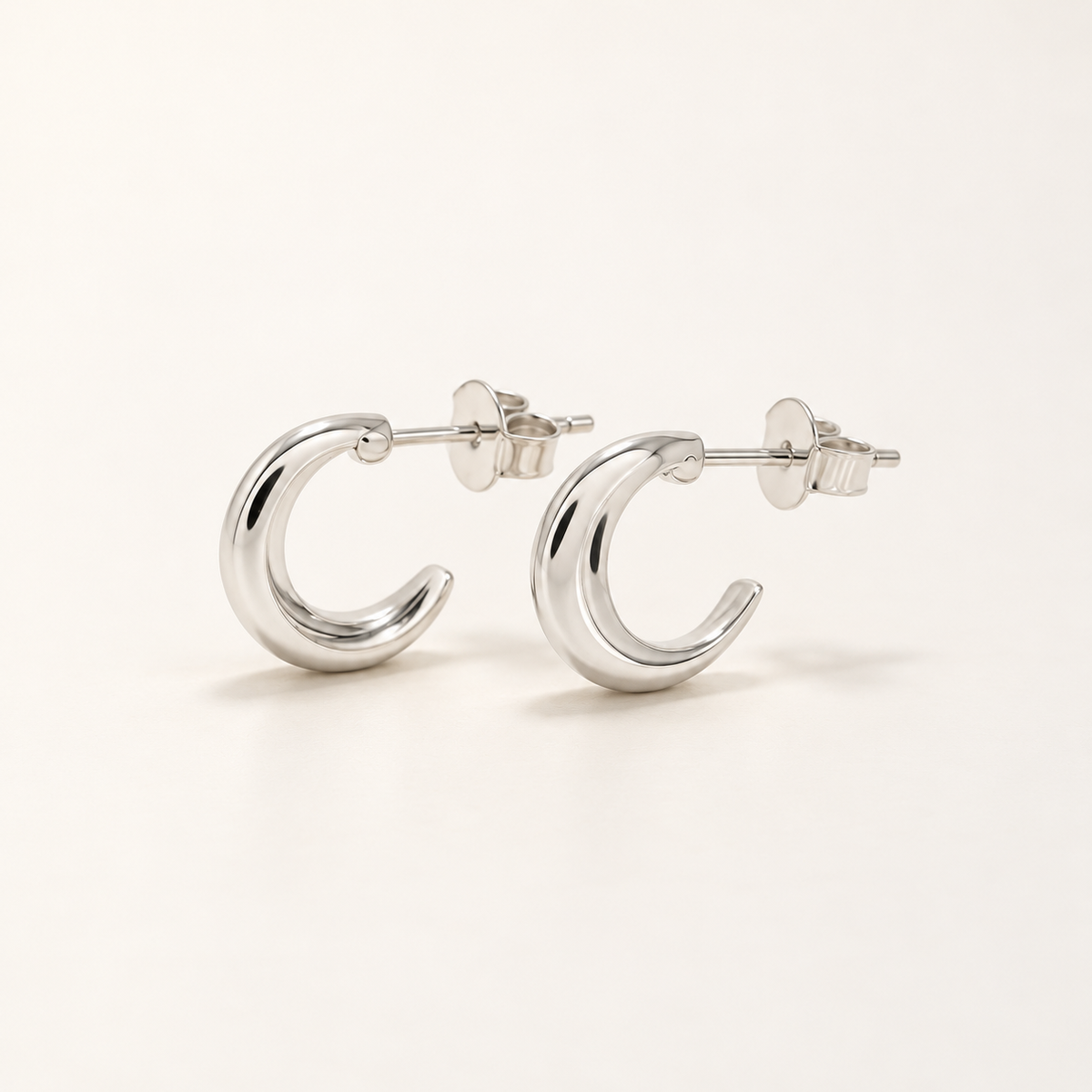 A pair of polished silver crescent hoop earrings with post and butterfly backings, displayed on a clean, light beige background.