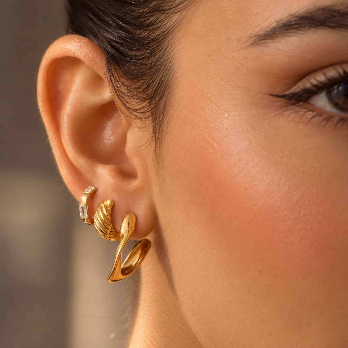 A close-up of a woman's ear wearing a stack of three gold huggie hoop earrings, including one with a small clear crystal accent.