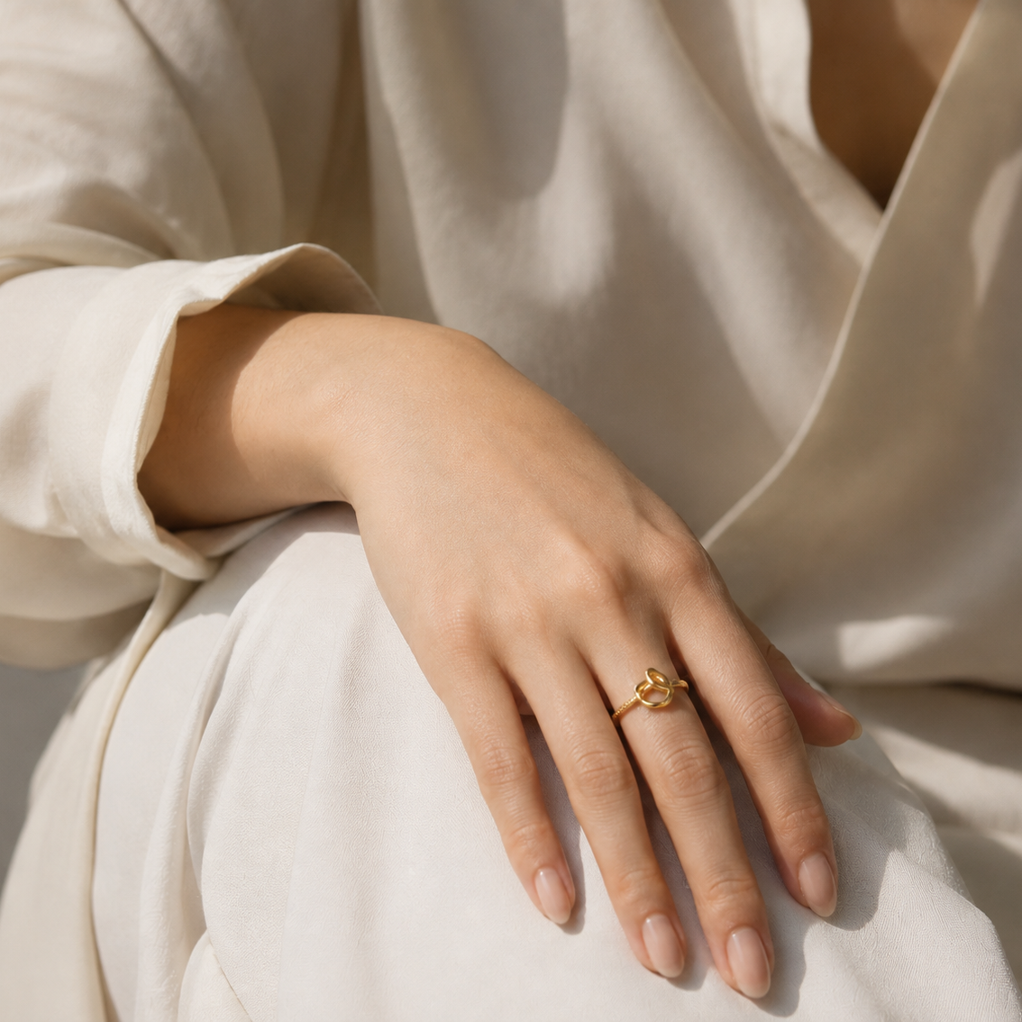 A close-up of a person's hand resting on their lap, wearing a delicate gold ring with a small floral or leaf-like motif. The person is wearing a soft, cream-colored blouse under warm, natural sunlight.