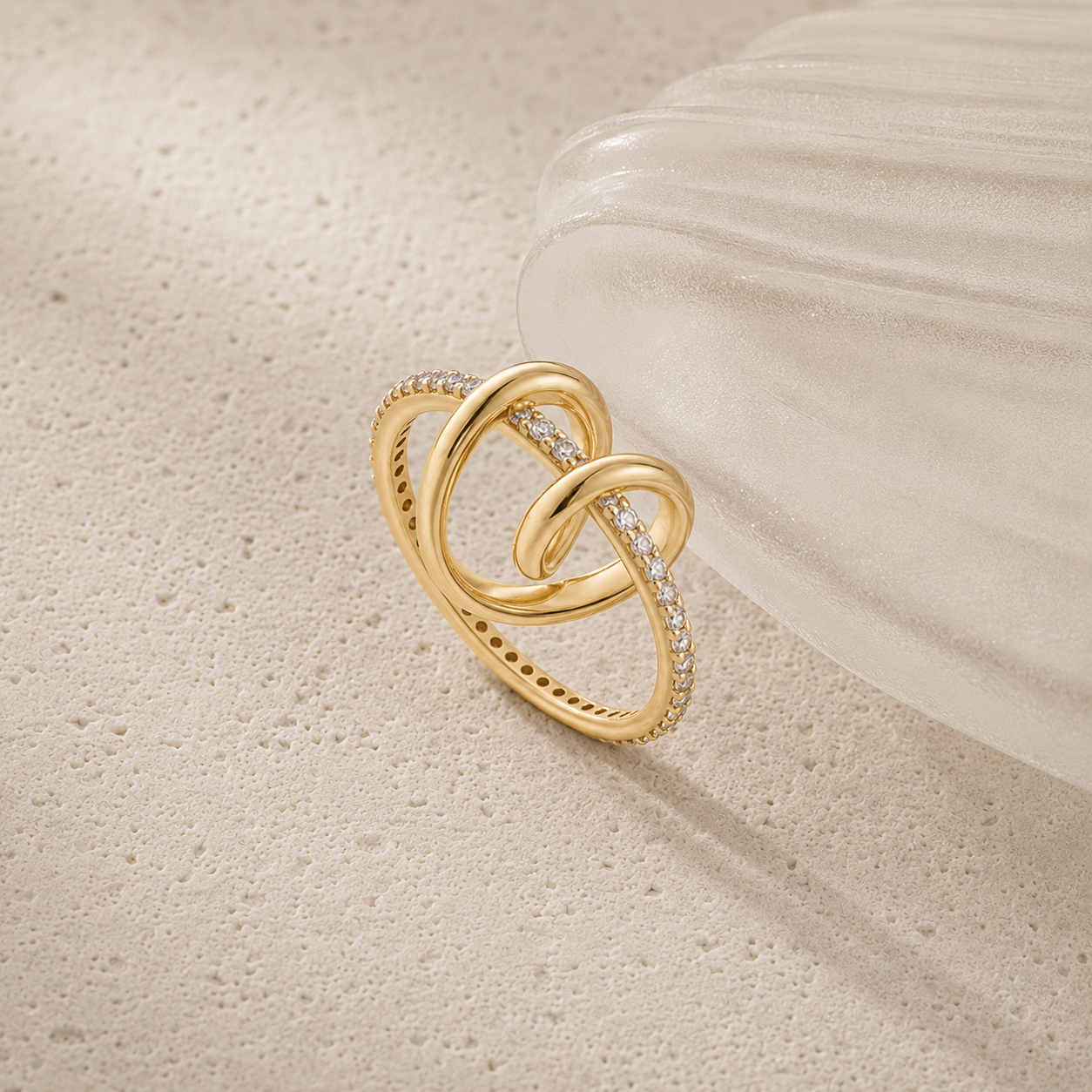 A gold interlocking knot ring with small clear stones set along one of the loops, displayed on a textured beige surface next to a translucent white shell.