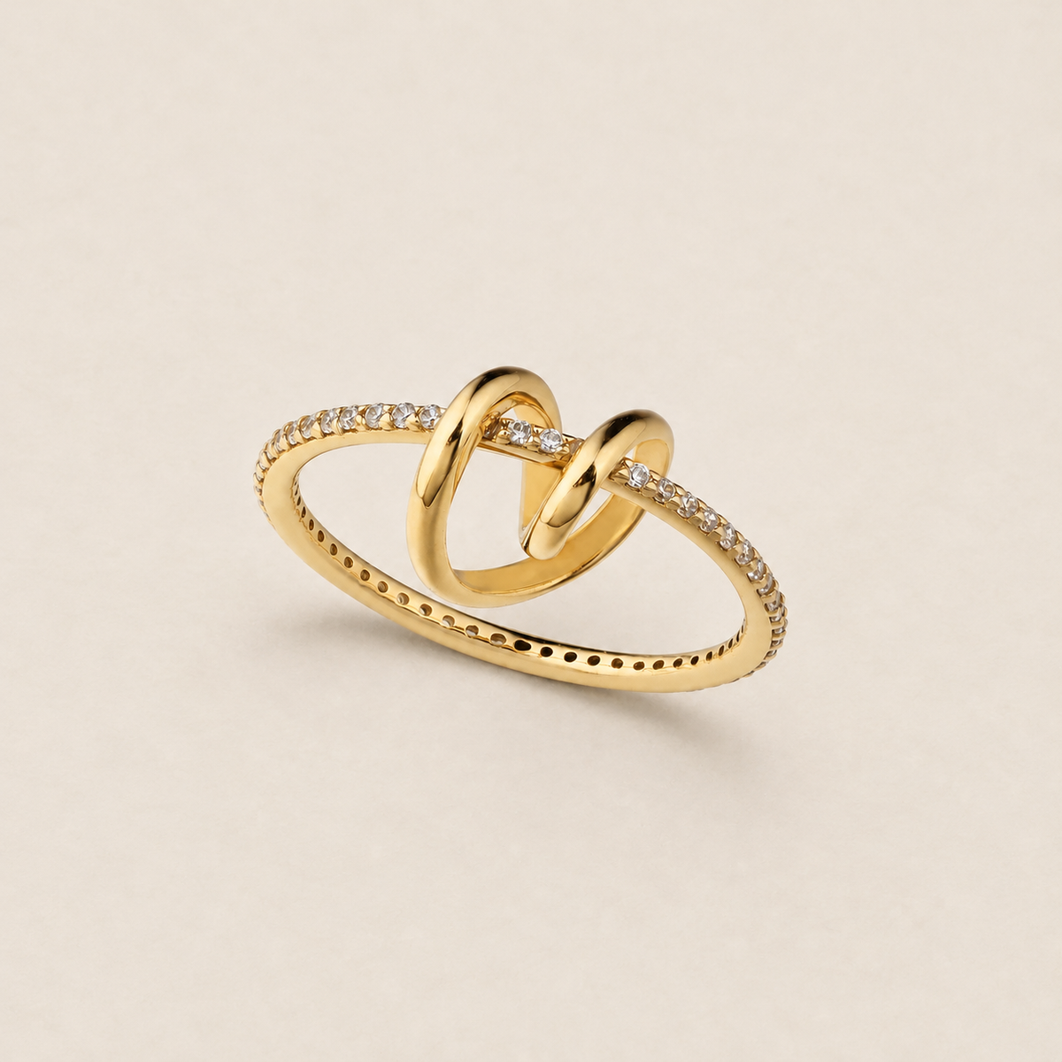 A gold ring featuring two interlocking knot-like loops, with pavé-set clear stones along the band and a row of small perforations on the lower band, photographed on a light beige background.