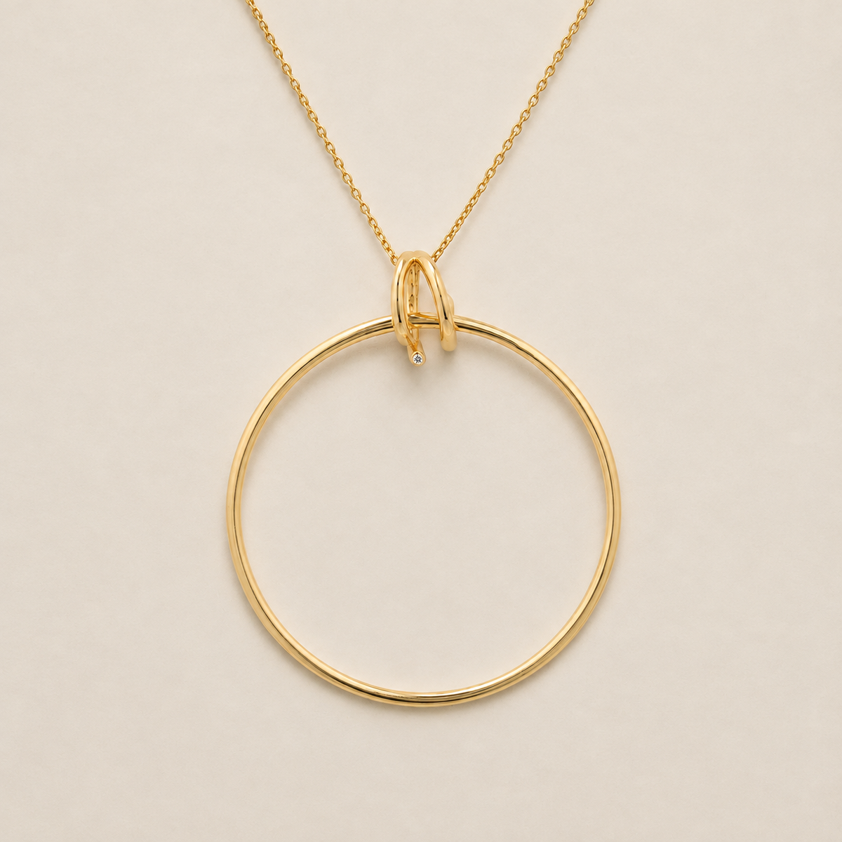 A gold necklace featuring a large, minimalist open circular pendant with a small, delicate interlocking loop at the top and a tiny clear gemstone accent, shown on a plain light beige background.