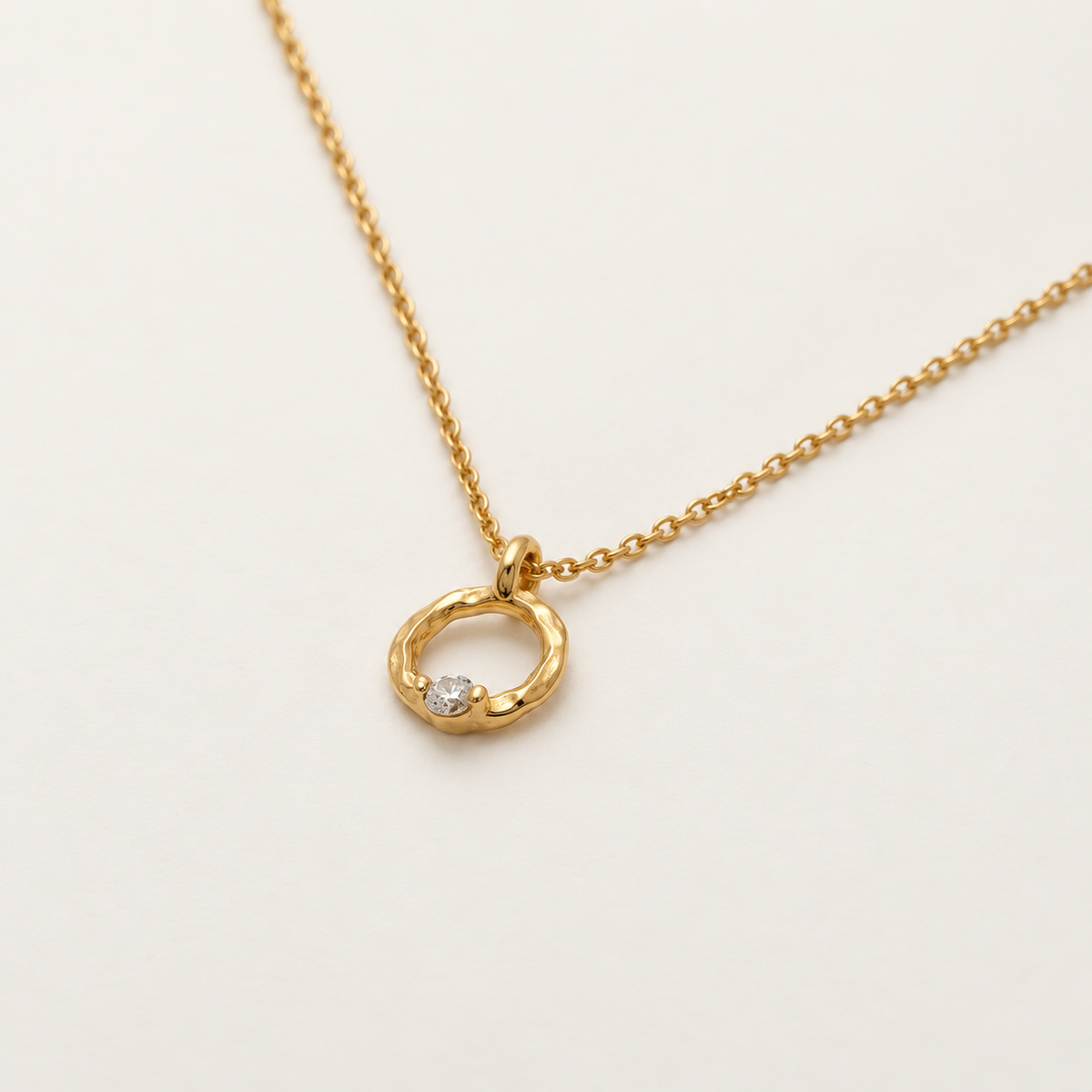 A gold necklace with a textured open ring pendant featuring a small clear gemstone, displayed on a clean off-white background.