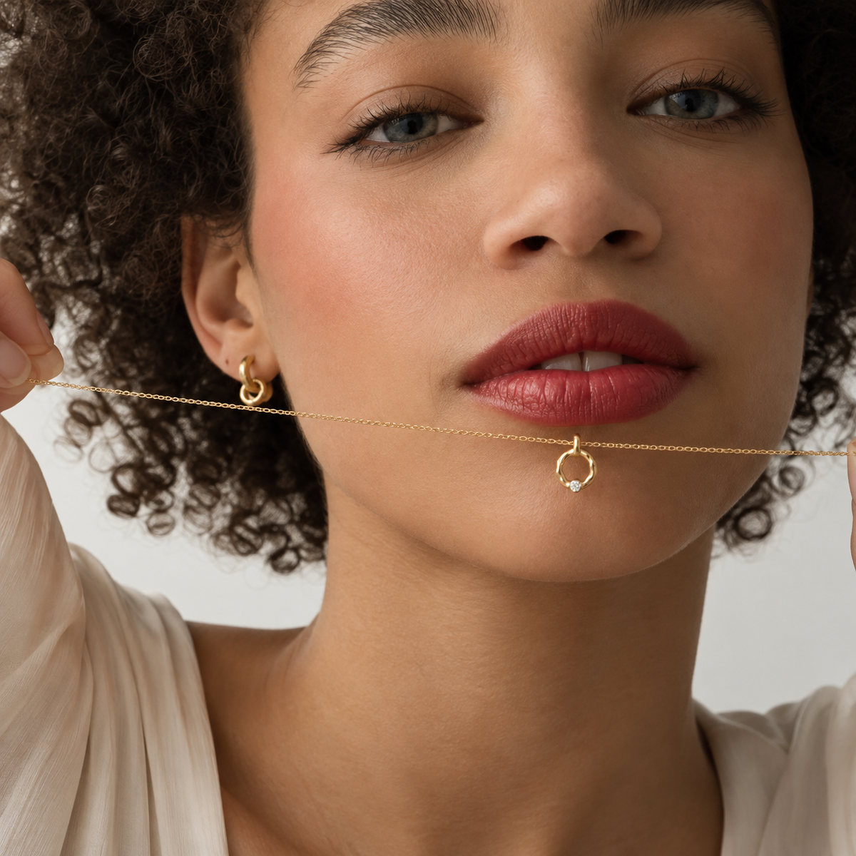 A close-up lifestyle shot of a woman wearing a delicate gold chain necklace with a small open hoop pendant, holding the chain with both hands against a clean, light background.
