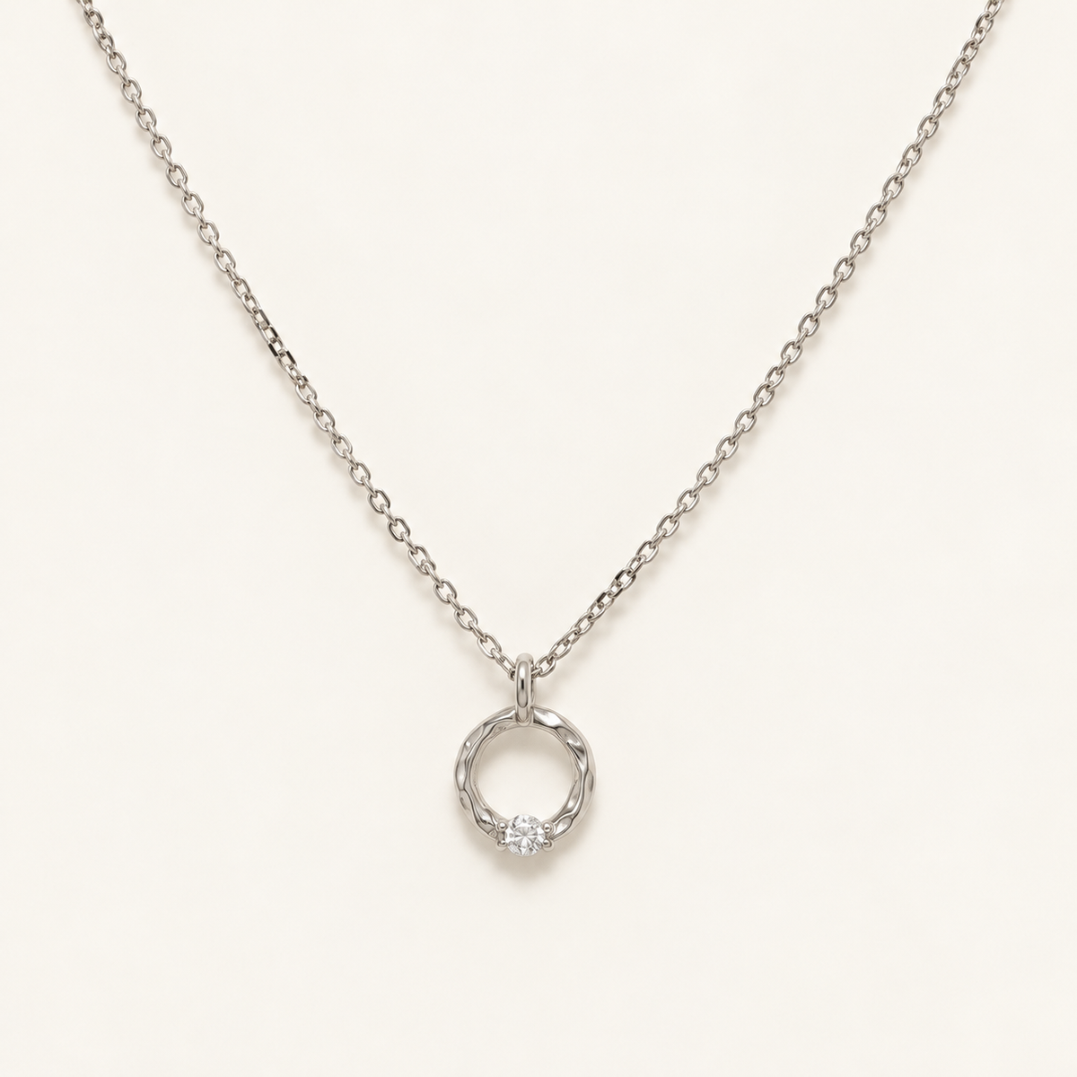 A delicate silver chain necklace with a small twisted circular pendant set with a single clear gemstone, displayed on a clean off-white background.