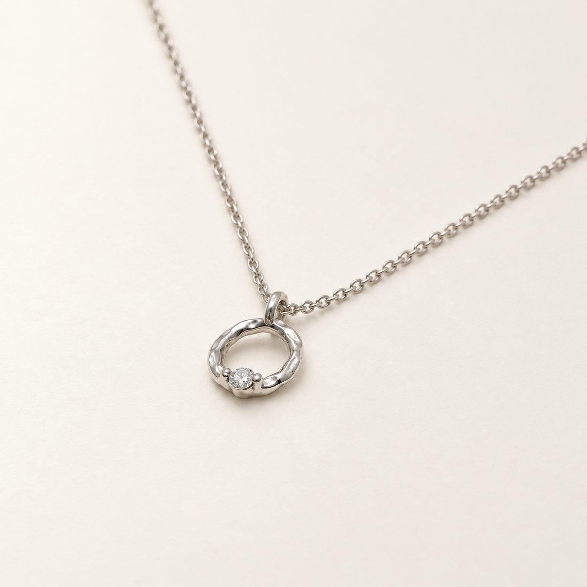 A silver chain necklace with a small twisted ring pendant featuring a single clear gemstone, displayed on a light cream background.