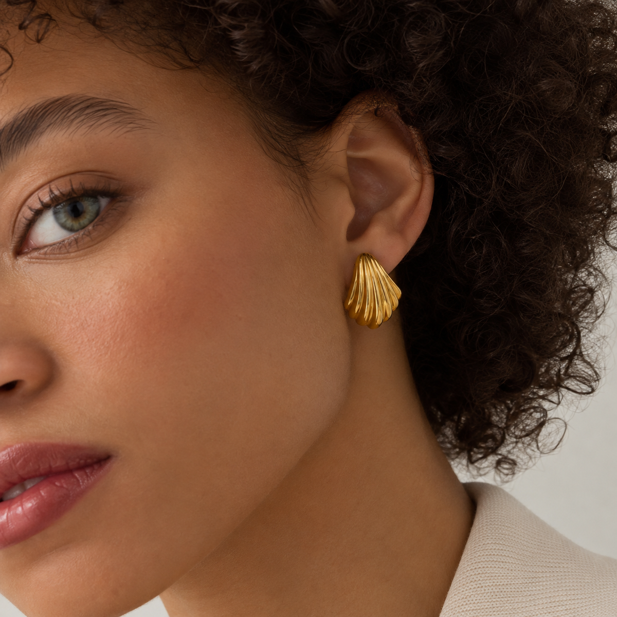 A close-up of a model wearing a textured gold shell-shaped stud earring, showing the ear and side of the face against a clean light background.