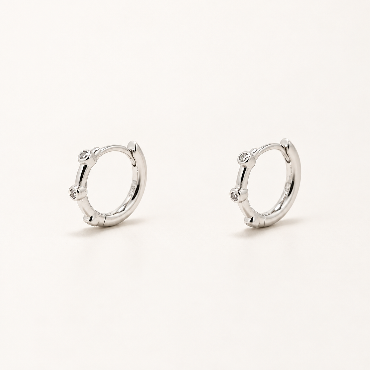 A pair of silver huggie hoop earrings with small clear gemstone accents on the front, displayed on a clean off-white background.