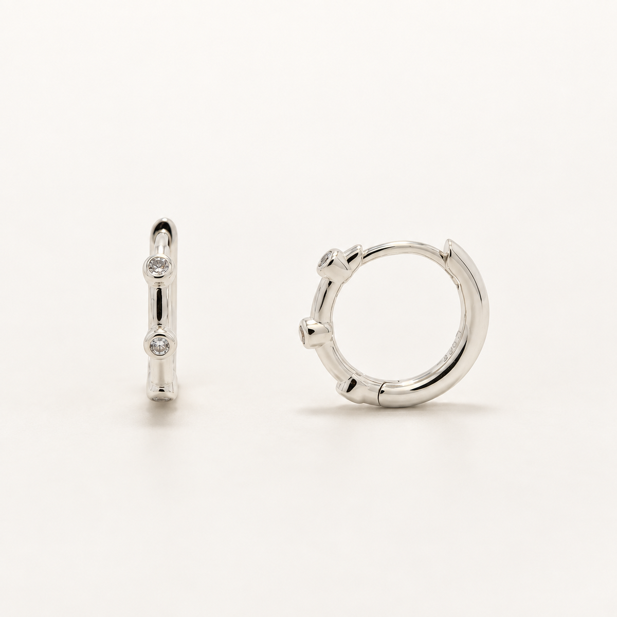 A pair of polished silver huggie hoop earrings, one shown from a side profile view with small clear stones and the other from a front-three-quarter view, set against a clean white background.