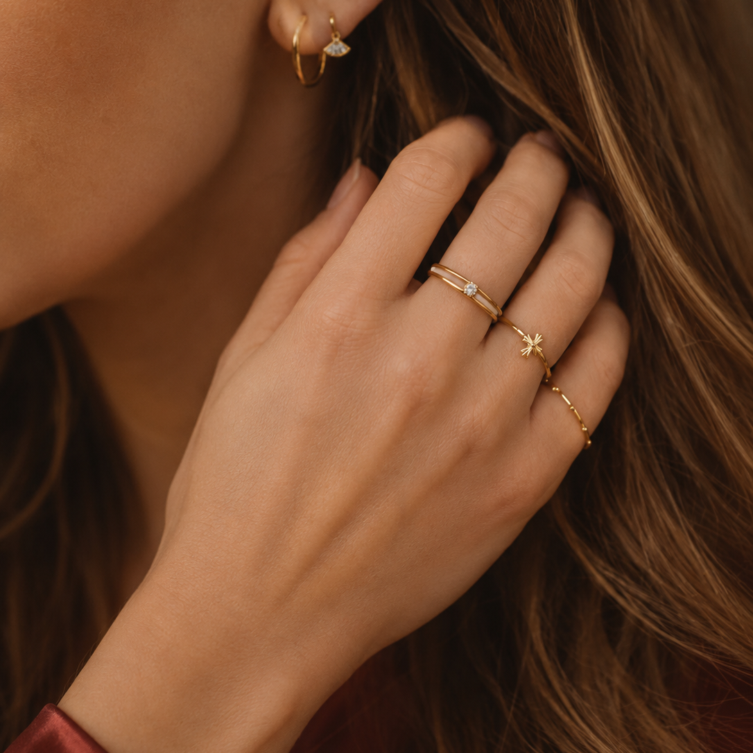 Close-up of a woman's hand and ear wearing delicate gold rings and hoop earrings, with her hair and a burgundy satin sleeve visible.