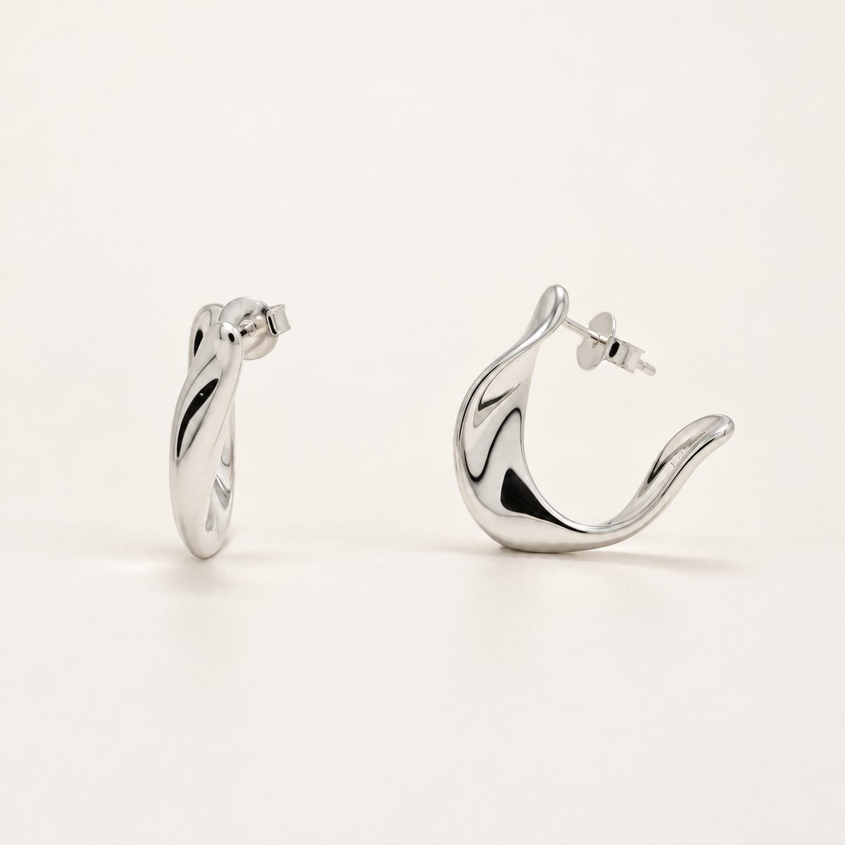 A pair of polished silver huggie hoop earrings with a fluid, organic wavy design, shown from two angles on a clean off-white background.