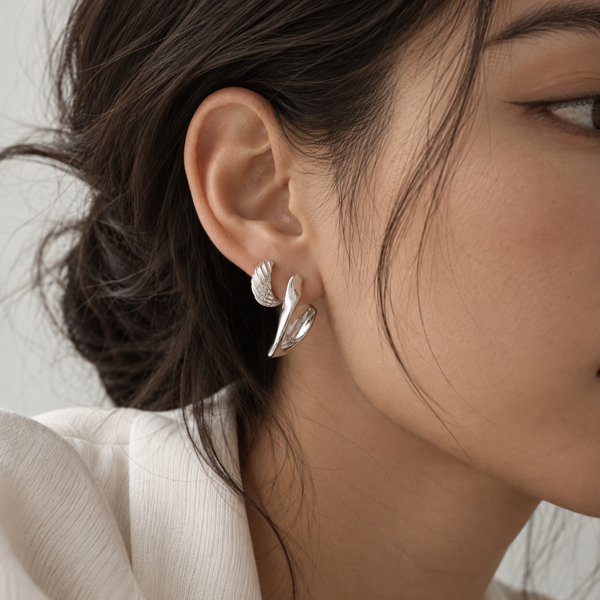 A close-up of a woman's ear wearing a pair of silver leaf-shaped stud earrings, with her dark hair and white top visible against a soft light gray background.