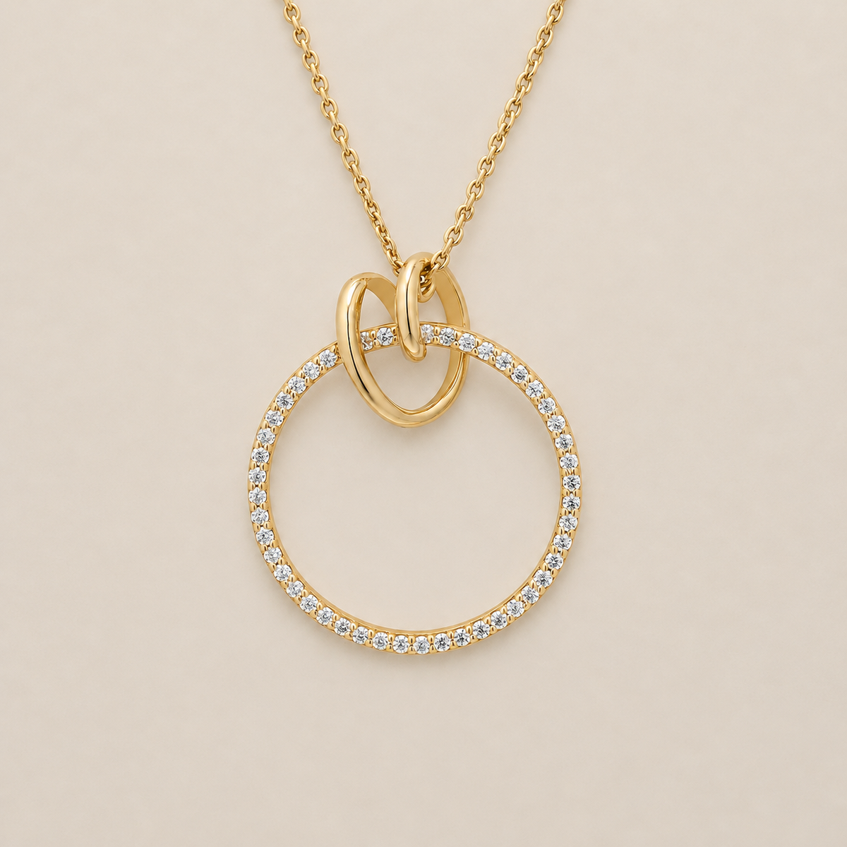 A gold pendant necklace featuring a large open circular ring paved with small clear stones, with two interlocking loop elements at the top and a delicate gold chain, shown on a light beige background.