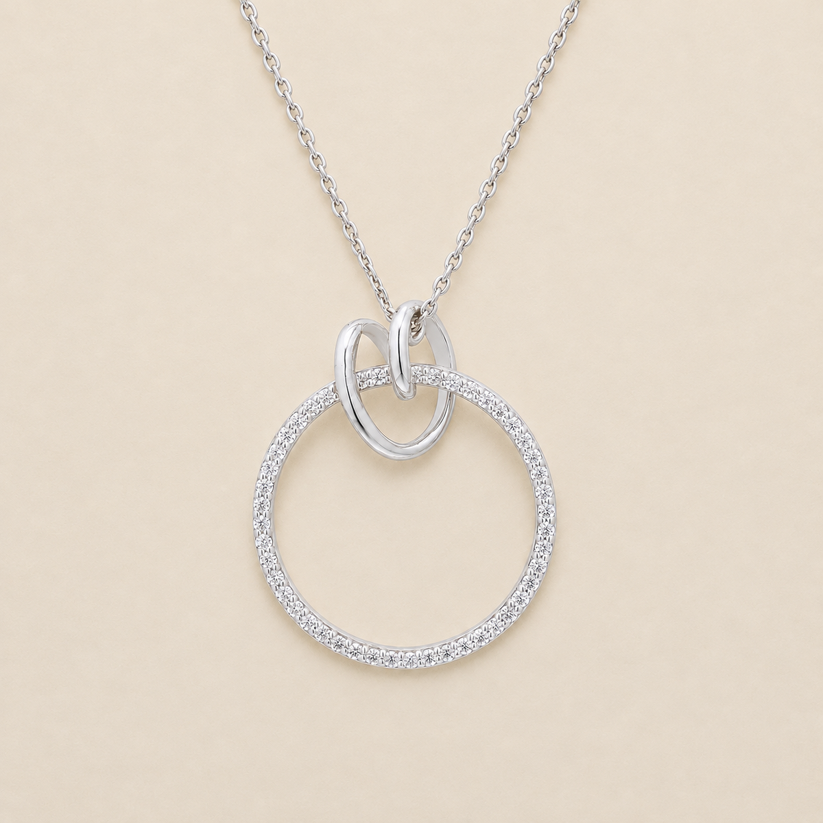 A silver necklace featuring a large open circle pendant paved with small clear stones, with a smaller polished oval loop at the top and a delicate chain, shown on a light beige background.